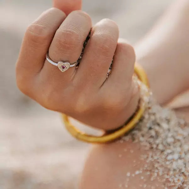 bague enfant petit coeur en argent avec une étoile sertie d'un zircon rose dans un coeur gravé portée sur une main qui tiens du sable