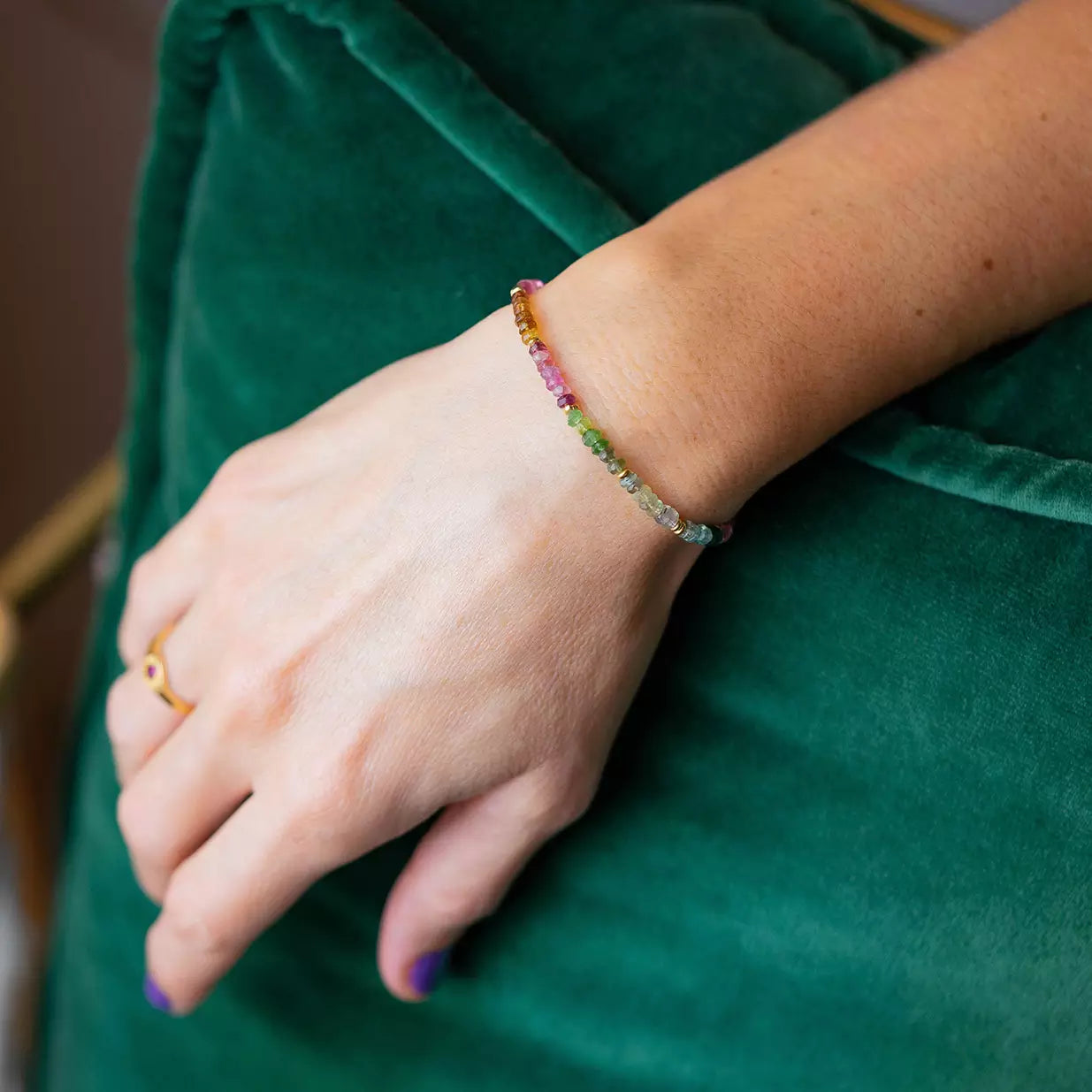 bracelet femme fait de pierres de tourmaline roses vertes et bleues portée sur une main aux ongles violet reposant sur un coussin vert