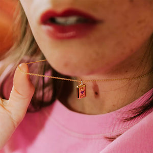 collier femme collection l'amoureuse plaqué or de forme rectangulaire serti de zircons couleur cristal et un zircon couleur fuschia en forme de coeur avec des gravures en forme de feuilles porté au cou par une femme avec du rouge à lèvre