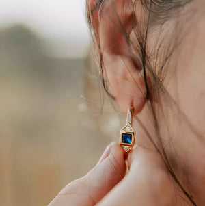 boucle d'oreille créole femme plaquée or sertie d'un zircon couleur bleu nuit et encadré de deux zircons couleur cristal portée sur une oreille par une femme qui la touche de son index et de son pouce
