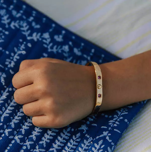 Bracelet jonc enfant plaqué or à gravure personnalisable avec un coeur au centre encadré de chaque côté par des gravures d'étoiles serties de pierres de zircons couleur fuchsia posé sur un tissu bleu 